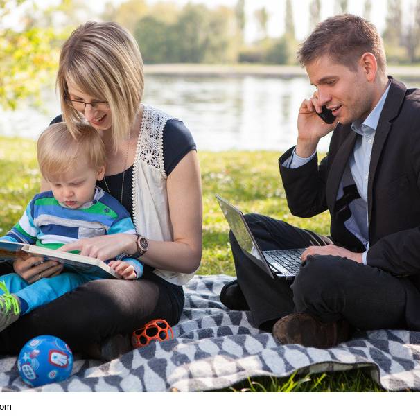 Eine junge Familie picknickt draußen in der Natur, während der Mann Büroarbeiten am Laptop erledigt.