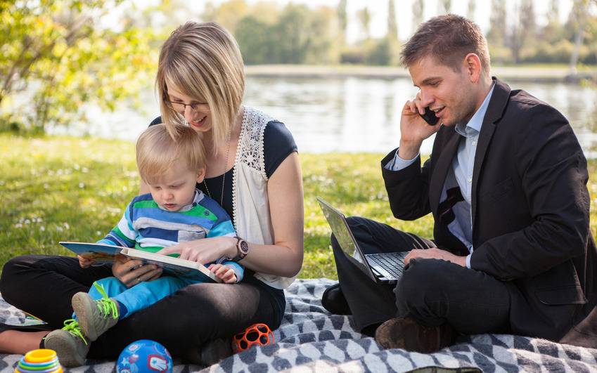Eine junge Familie picknickt draußen in der Natur, während der Mann Büroarbeiten am Laptop erledigt.