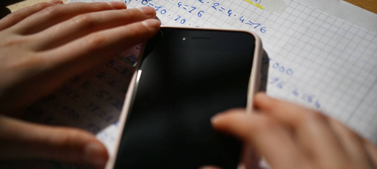 Ein Kind hält ein Smartphone über einem Blatt mit Mathematikaufgaben - das Gerät liegt auf dem Arbeitsblatt.